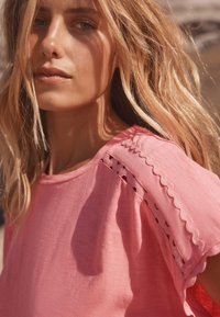 Rózsaszín rövid ujjú felső hullámos szegéllyel és csipkés részletekkel a vállaknál, könnyű anyagból készült, kényelmes szabással.