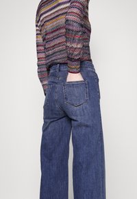 Personne portant un jean bleu à jambes larges, une main dans la poche arrière et un haut à manches longues à motif zigzag multicolore
