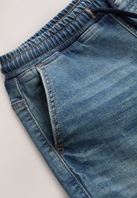 Close-up van blauwe spijkerbroek met elastische tailleband en schuin geplaatste voorzak met zichtbare stiksels.