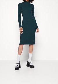 Teallista raidoitettu midi mekko pitkillä hihoilla ja korkealla kaula-aukolla. Vaatteena mustat korkeat kengät ja valkoiset sukat. Minimalistinen muotoilu, tiukka istuvuus.