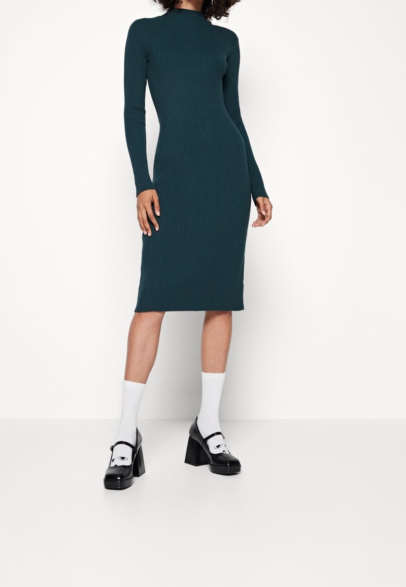 Teallista raidoitettu midi mekko pitkillä hihoilla ja korkealla kaula-aukolla. Vaatteena mustat korkeat kengät ja valkoiset sukat. Minimalistinen muotoilu, tiukka istuvuus.