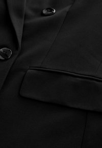 Sort skræddersyet blazer med en glat tekstur, to sorte knapper og en enkelt welt-lomme. Syede detaljer langs kanterne.