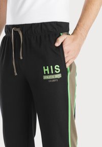 Schwarze Sporthose mit grünen und braunen Seitestreifen. Verfügt über einen elastischen Bund und ein aufgedrucktes "H.I.S ATHLETIC DEPT. CHAMPS" Logo.