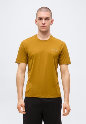 Fiatal férfi borotvált fejjel, mustár sárga rövid ujjú ingben és fekete nadrágban, egy egyszínű világos háttér előtt áll.