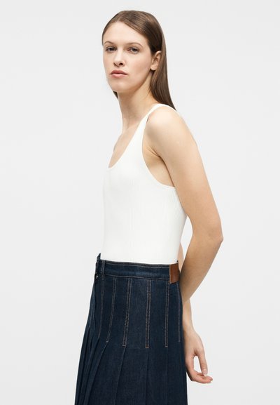 Γυναίκα με ίσια καστανά μαλλιά, ντυμένη με λευκή μπλούζα χωρίς μανίκια και σκουρό μπλε πλισέ φούστα, στέκεται πλευρικά μπροστά σε ένα απλό λευκό φόντο.