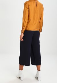 Sininen satiinipaita, jossa on pitkät hihat ja takana avainreikäyksityiskohta, yhdistettynä leveälahkeisiin tummansinisiin culotteihin ja valkoisiin lohkare-korkokenkiin.