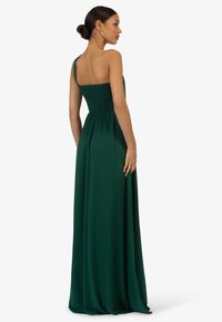 Langes, ärmelloses, dunkelgrünes Kleid mit einem gerüschten Oberteil und fließendem Rock. Es verfügt über einen einzelnen Schulterträger und eine glatte Stofftextur.