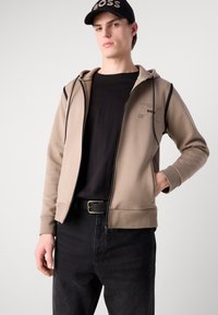 Veste à zip beige avec des accents noirs, texture douce, poches kangourou. Portée sur une chemise noire et associée à un jean foncé et une casquette noire.