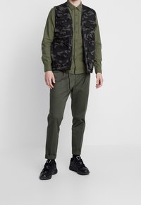 Zwarte camouflageshirt met meerdere zakken, groen overhemd met knopen, olijfgroene broek en zwarte sneakers; casual outfit met veelzijdige materialen.