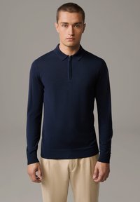 Navyblaues Langarm-Polohemd mit einem Halbzipper-Kragen, aus einem glatten Stoff gefertigt, kombiniert mit hellbeigen trousers. Einfaches und figurbetontes Design.