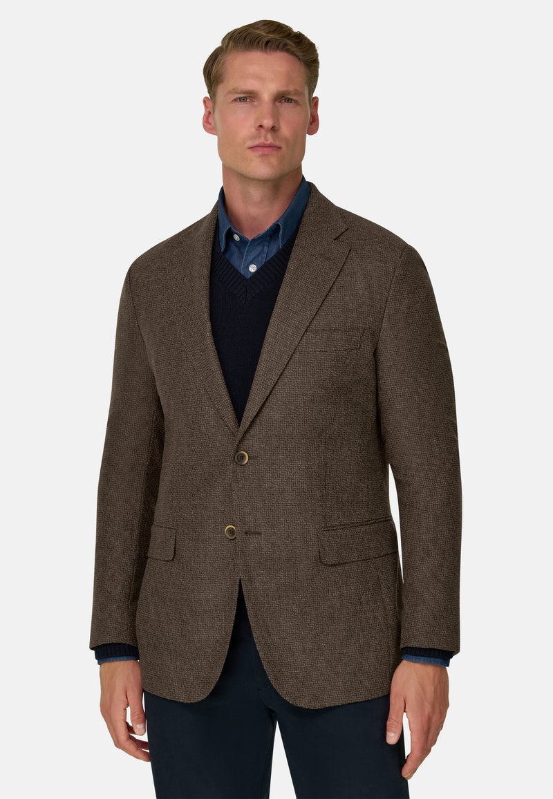 Homme portant un blazer marron texturé par-dessus un pull bleu marine et une chemise bleue à col, sur un fond clair uni.