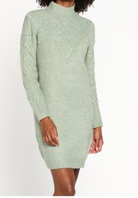 Robe pull en tricot à manches longues de couleur vert clair. Présente un col montant et un motif en losanges texturé sur la poitrine avec des poignets et un ourlet côtelés.