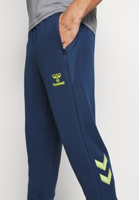 Person trägt dunkelblaue Jogginghose mit gelbem hummel-Logo und Chevron-Design, Hand steckt in der Tasche, graues Hemd teilweise sichtbar.