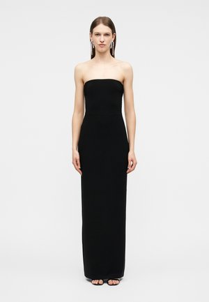 Kvinne iført en svart stroppeløs kjole som går helt ned til gulvet, med svarte sandaler med åpen tå og lange dråpeformede øredobber, stående foran en hvit bakgrunn.