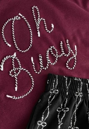 Material maro cu broderie albă în stil funie care formează cuvintele „Oh Buoy”. Pantaloni scurți negri cu model alb de noduri și talie elastică dedesubt.