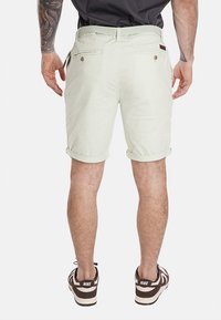 Pantaloni corti verde chiaro con risvolti, tasche posteriori e una cintura intrecciata testurizzata. Indossati con sneakers Nike marroni e bianche.