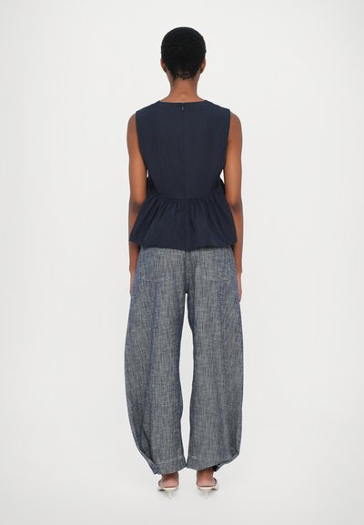 Jūras spēku krāsas bez piedurknēm peplum tops, kombinēts ar brīvām, tekstūrētām zilām džinsu biksēm un gaišiem, norādītiem apaviem.