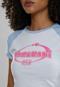 Camiseta blanca con mangas raglán en azul claro, que presenta un destacado logo rosa que dice "KARLKANI" y detalles de corazones juguetones. Textura suave.