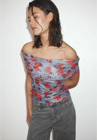 Donna con i capelli bagnati che indossa un top azzurro scollato con stampa floreale rossa e jeans neri a vita alta sbiaditi, in piedi davanti a uno sfondo bianco.