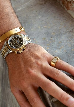 Gouden en zilveren polshorloge met zwarte wijzerplaat, chronograaf functies, geborsteld metalen band. Vergezeld door een gouden armband en ring met edelstenen.