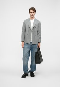 Νεαρός άνδρας που φορά γκρι διπλόχρωμο σακάκι, λευκό πουκάμισο, χαλαρές μπλέ τζιν παν trousers, μαύρα παπούτσια, κρατώντας μια μαύρη δερμάτινη τσάντα, σε στάση μπροστά από λευκό φόντο.