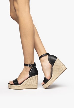 Bene iført sorte kilehælssandaler med ankelrem og fletninger i jute på plateauhælene samt sølvnitter bagpå.