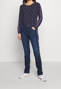 Tmavě modrý top s dlouhým rukávem, zdobený víc barevným vzorem a pruhovanými akcenty na rukávech, v kombinaci s tmavě modrými džínami s bočním pruhem a bílými teniskami.