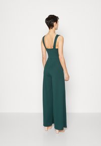 Combinaison verte à jambes larges, au dos nu, avec des bretelles ajustables et un corsage ajusté. Tissu lisse avec une texture subtile.