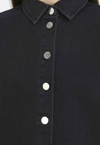 Camicia in denim scuro con colletto classico, bottoni a pressione argentati sul davanti e un tessuto a trama liscia.