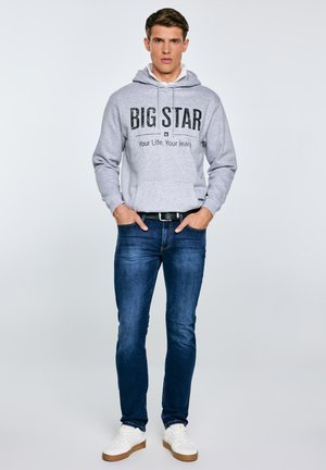 Mladý muž stojící s rukama v kapse, oblečený v šedé mikině s nápisem „BIG STAR“, modrých džínách, černém pásku a bílých teniskách.