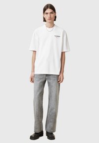 White cotton t-shirt with short sleeves, featuring a small black logo. Worn with faded grey straight-leg jeans and black boots.
