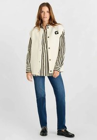 Roomkleurig geribde tanktop met zwarte knopen, gecombineerd met een zwart-wit gestreept shirt en een blauwe spijkerbroek. Zwarte loafers maken de outfit compleet.