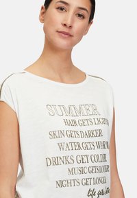 T-shirt blanc à manches courtes avec un texte doré métallique sur le thème de l'été. Tissu doux, coupe décontractée, col rond et design simple.