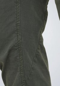 Olivgrüne Hosen mit glatter Textur, die diagonale Nähten und kontrastierenden Stichen an den Seiten für strukturelles Interesse aufweisen.