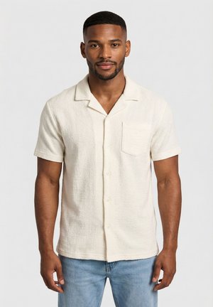 Uomo in piedi, indossa una camicia beige a maniche corte con texture, bottoni e taschino sul petto, abbinata a jeans azzurri, su uno sfondo semplice.