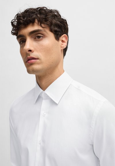 Chemise blanche à boutons avec un col classique, texture lisse et coupe ajustée. Présente une patte de boutonnage et des épaules structurées.