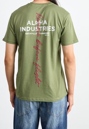 Olivengrøn bomulds-T-shirt med teksten "ALPHA INDUSTRIES" og "Remind Before Flight" i hvidt og mørkerødt på ryggen. Korte ærmer.