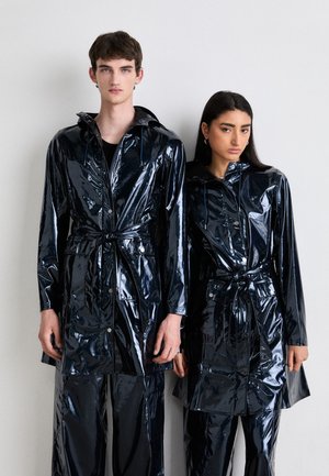 Black, glossy vinyl raincoats with long sleeves, drawstrings, and belt details, worn over matching pants, displayed on two individuals.