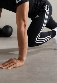 Svarta träningsbyxor har vit tre-stripes detaljer längs sidorna. Tyget verkar slätt och elastiskt, vilket gör dem lämpliga för träning.