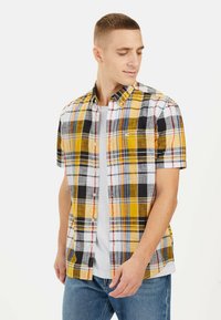 Kortemouwen geruit shirt met een geel, wit, zwart en rood patroon, knoopsluiting en een casual pasvorm. Gekozen over een wit t-shirt.