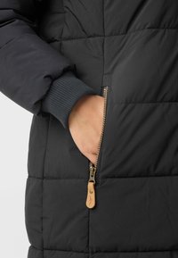Hand resting inside the partially unzipped pocket of a black padded jacket with ribbed cuff and leather zipper pull tab.
