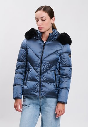 Frau trägt eine glänzende blaue gesteppte Jacke mit schwarzen Pelzakzenten auf den Schultern und hellblaue Jeans, stehend vor einem schlichten Hintergrund.