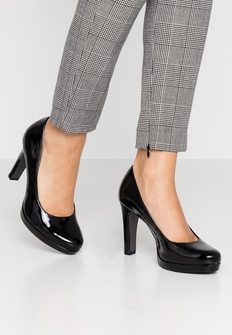 Personne portant des escarpins en cuir verni noir et un pantalon à carreaux gris, debout sur un sol clair devant un fond blanc.