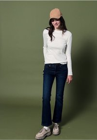 Hvid langærmet skjorte, mørkeblå jeans, beige kasket og lysebeige sko, stående foran en grøn baggrund.