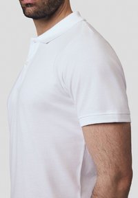 Biała koszulka polo wykonana z bawełny, z kołnierzykiem, dwoma guzikami, krótkimi rękawami i fakturowanym wzorem materiału. Pokazana w widoku z boku.