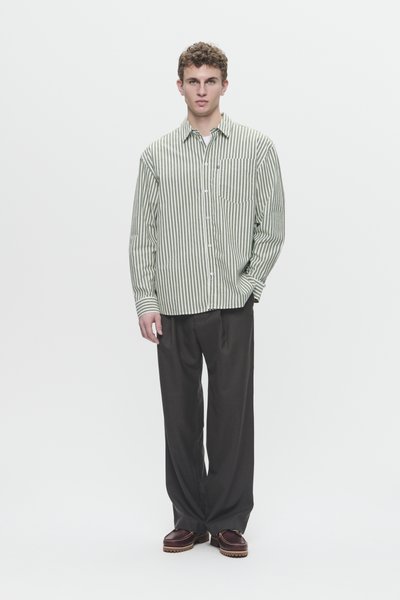 Giovane in piedi che indossa una camicia a righe verde e bianca con bottoni, pantaloni larghi scuri e scarpe marroni con suole spesse.
