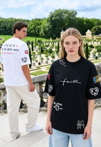 Jeune femme en noir et jeune homme en t-shirts graphiques blancs se tiennent à l'extérieur près d'une rambarde en pierre avec un jardin en arrière-plan.
