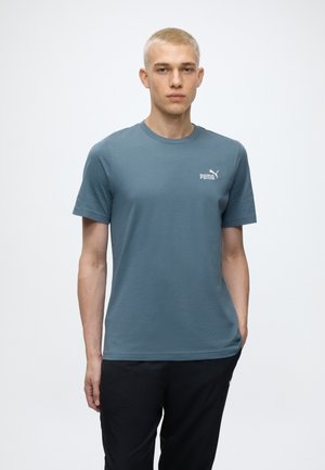 Jeune homme aux cheveux blonds courts portant un t-shirt bleu Puma et un pantalon noir sur un fond blanc uni.