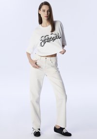 Weißes, kurz geschnittenes T-Shirt mit schwarzem "Floyd"-Schriftzug, kombiniert mit hoch taillierten weißen gerade geschnittenen Jeans und schwarzen Schuhen mit weißen Schnürsenkeln.