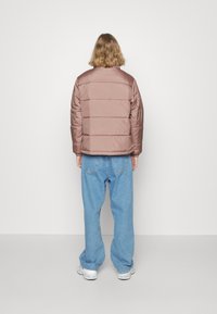 Giacca a vento marrone con cuciture orizzontali, abbinata a jeans larghi azzurri chiari e sneakers bianche. Tessuto liscio, design minimalista.